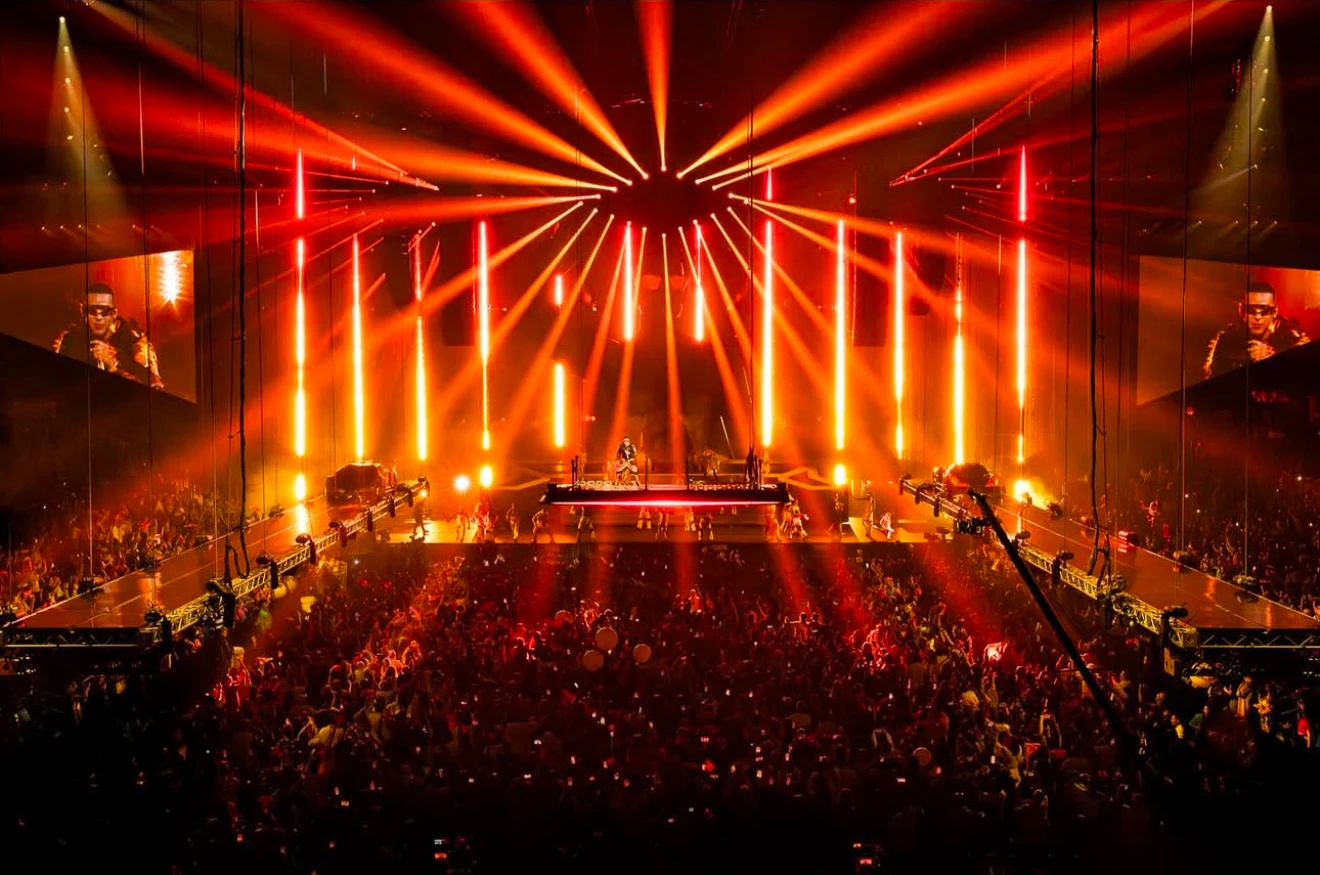 A wide arena stage filled with red light beams and large live video screens.